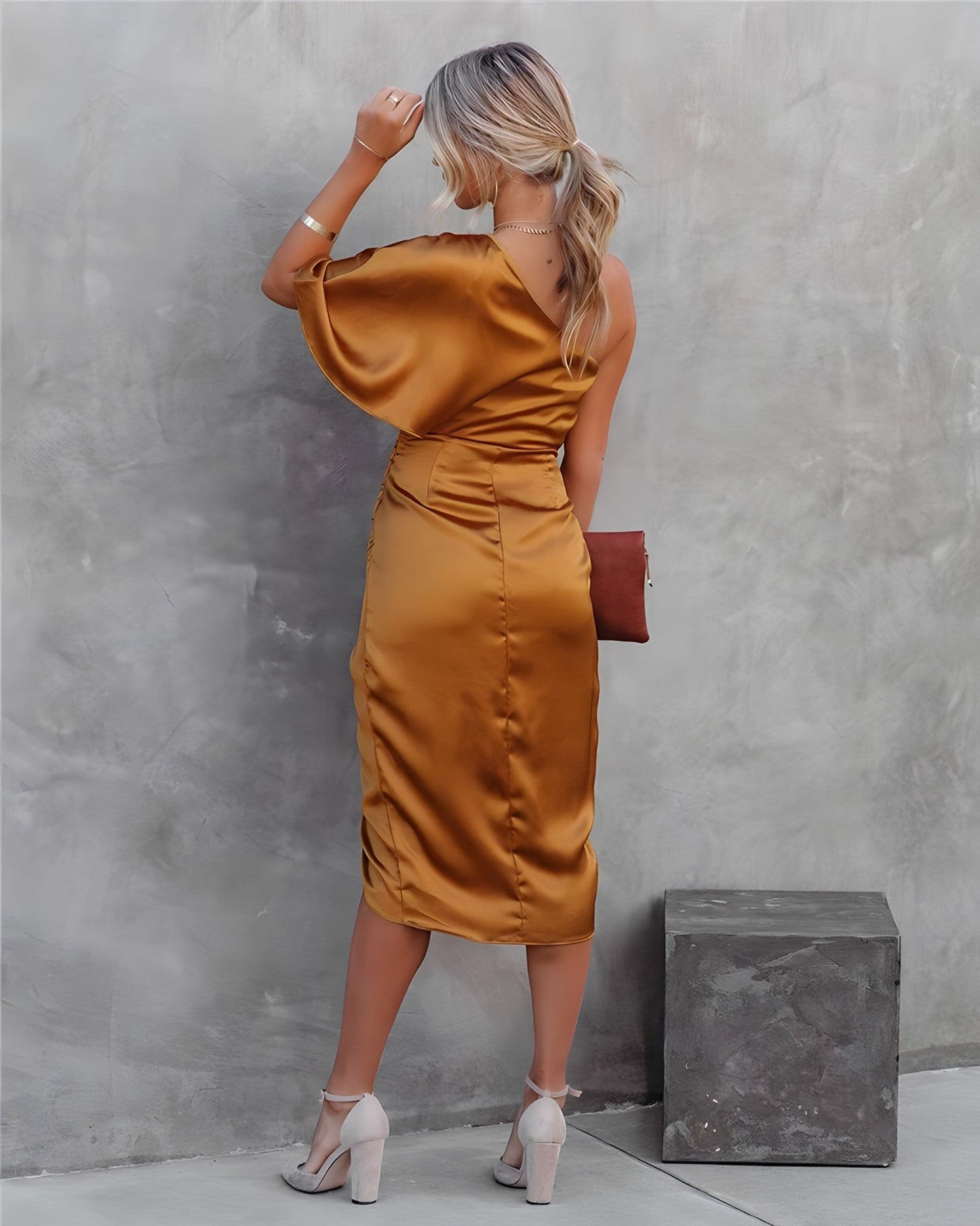 Femme en robe satinée dorée, dos tourné, tenant une pochette marron, posant contre un mur gris. Mode élégante, tenue de soirée chic.