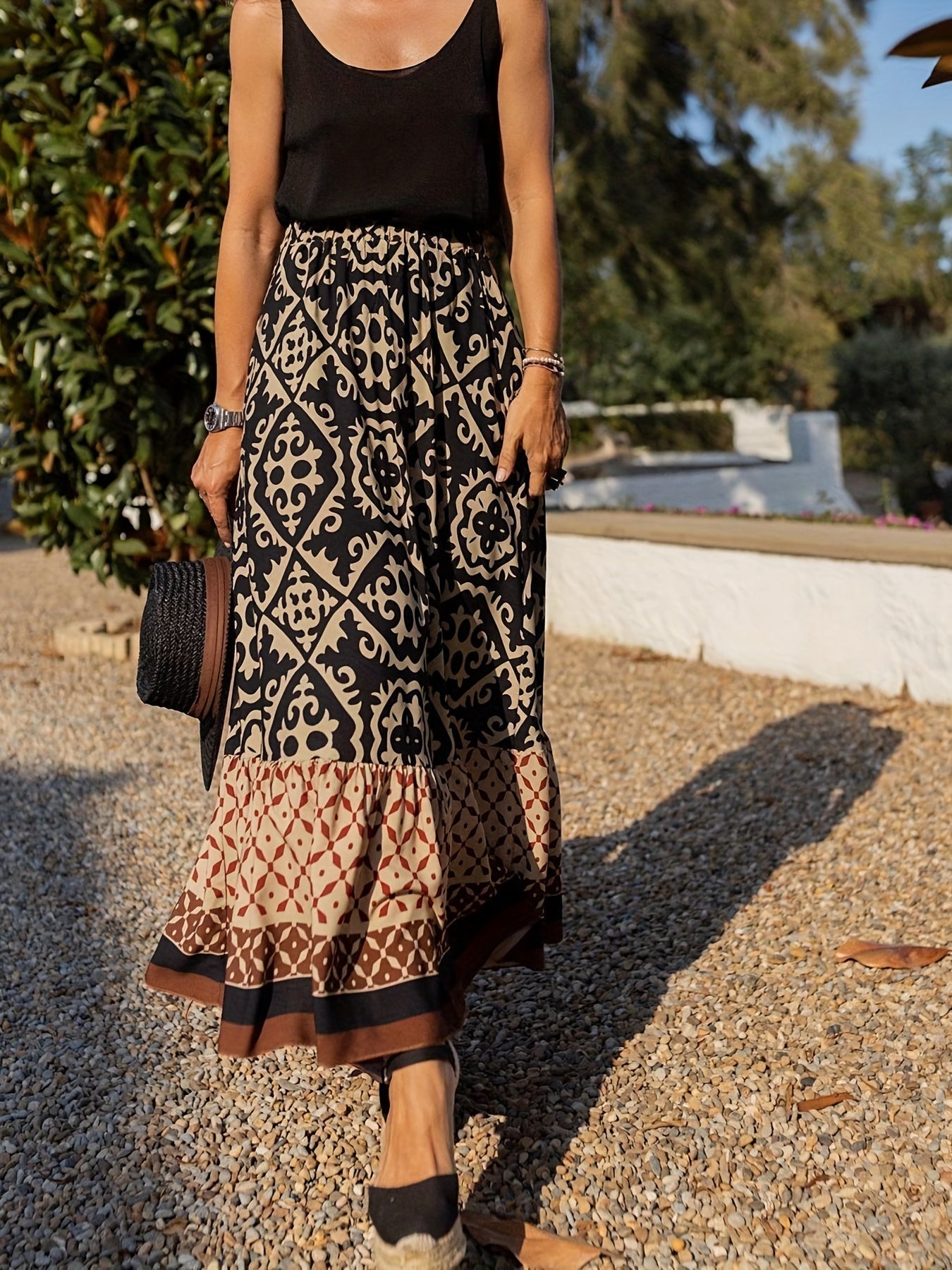 Femme en jupe longue bohème noire et beige à motifs géométriques, portant un débardeur noir et des espadrilles, marchant sur un chemin de gravier.