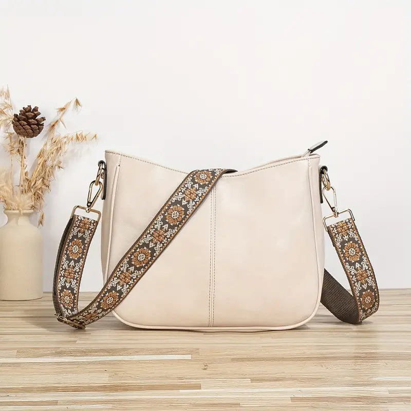 Sac à main beige en cuir avec bandoulière brodée florale, posé sur une table en bois, vase décoratif à côté. Accessoire mode élégant et tendance.