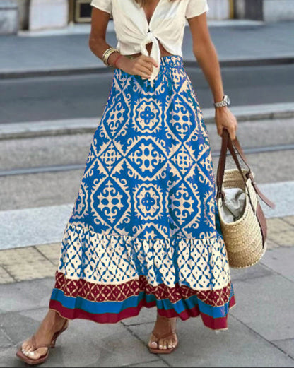 Femme portant une jupe longue bohème bleue à motifs géométriques, chemisier blanc noué, sandales, sac en osier, marchant en ville.