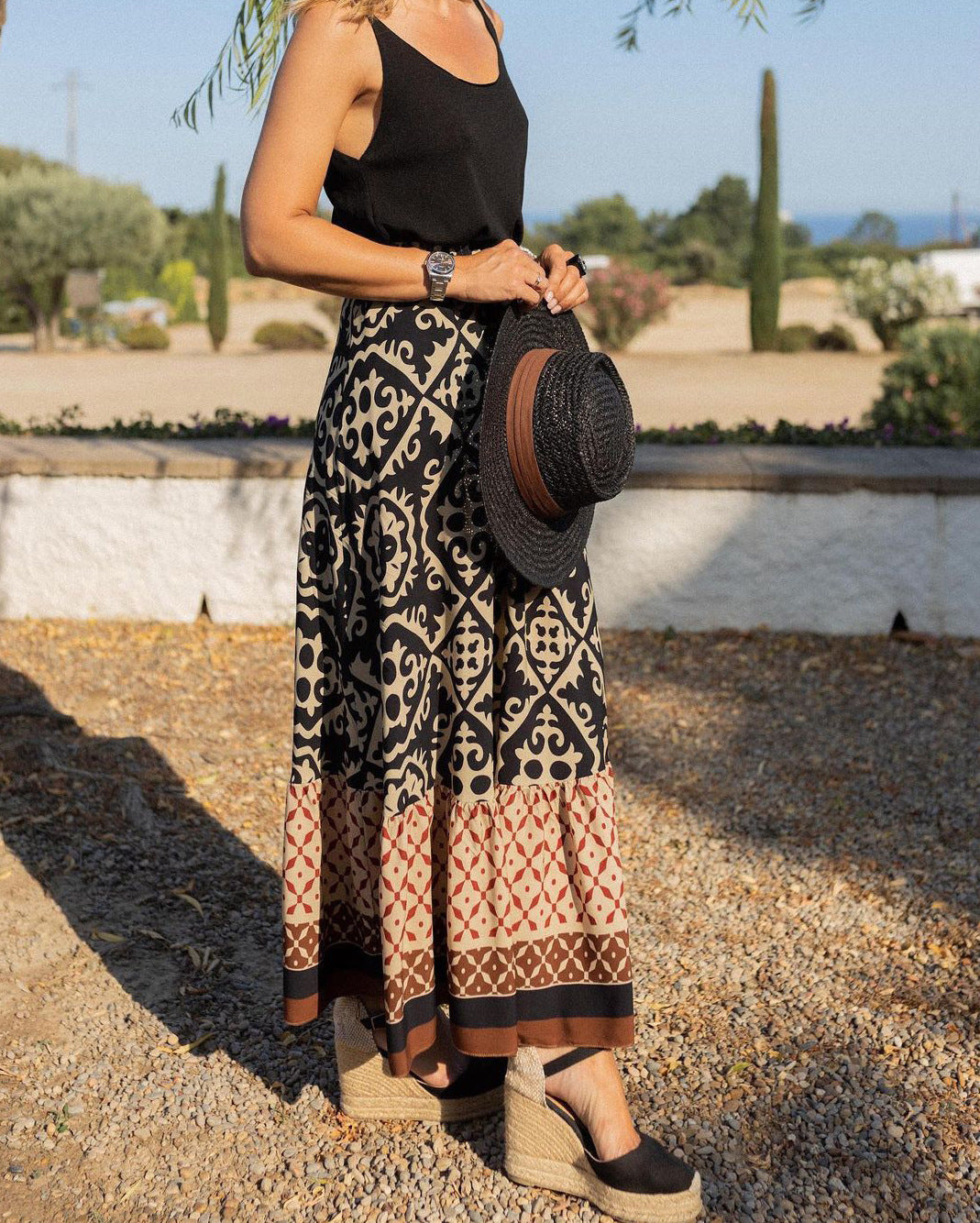 Femme en jupe longue bohème à motifs géométriques, portant un chapeau noir et des espadrilles compensées, dans un jardin méditerranéen ensoleillé.