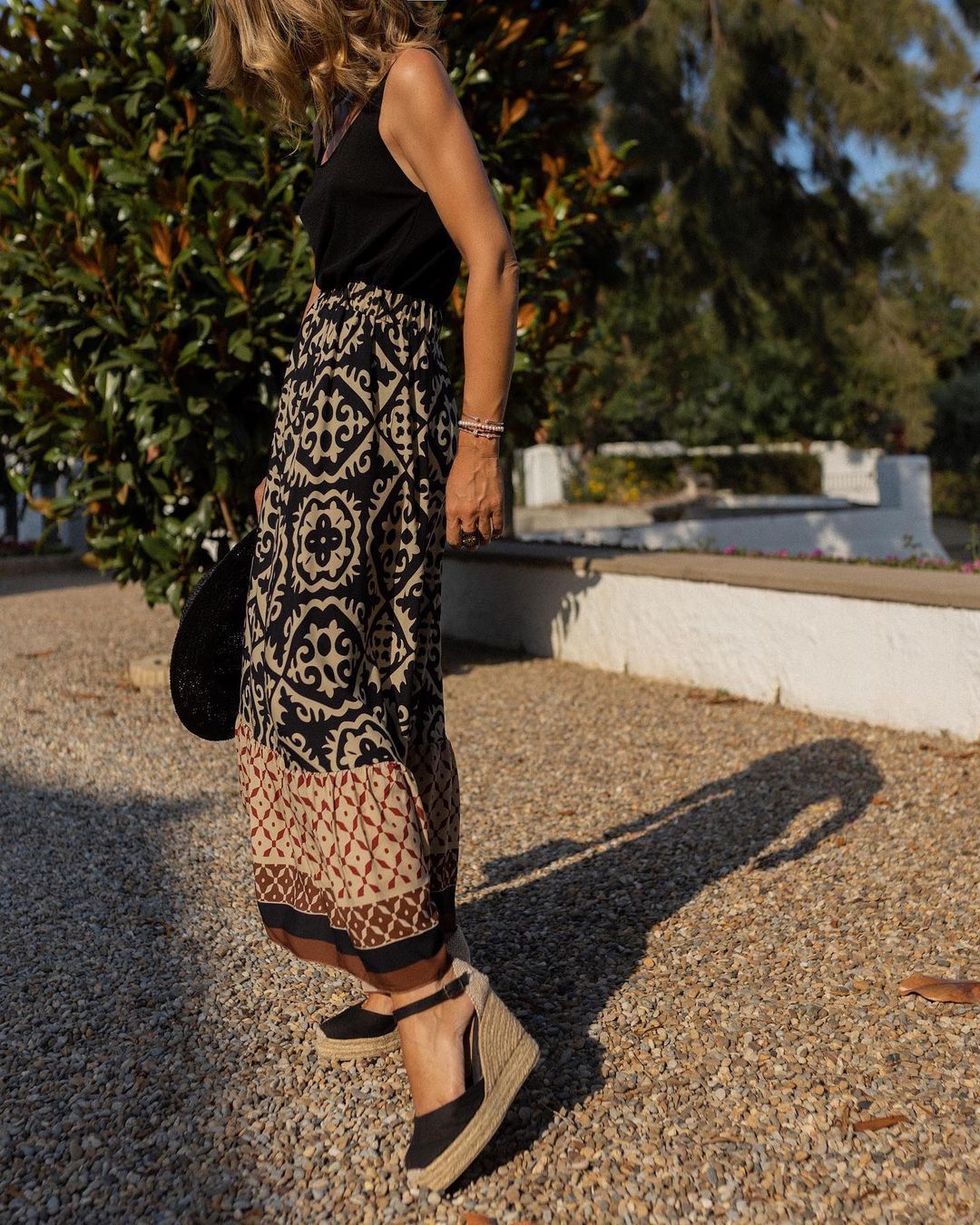 Femme en robe longue à motifs géométriques noirs et beiges, portant des espadrilles compensées noires, marchant sur un chemin de gravier ensoleillé.