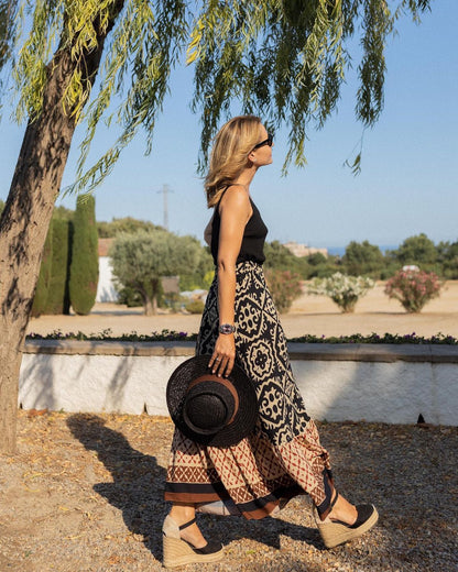 Femme marchant sous un arbre, portant une jupe longue à motifs et un chapeau noir, par une journée ensoleillée. Mode estivale, style bohème.