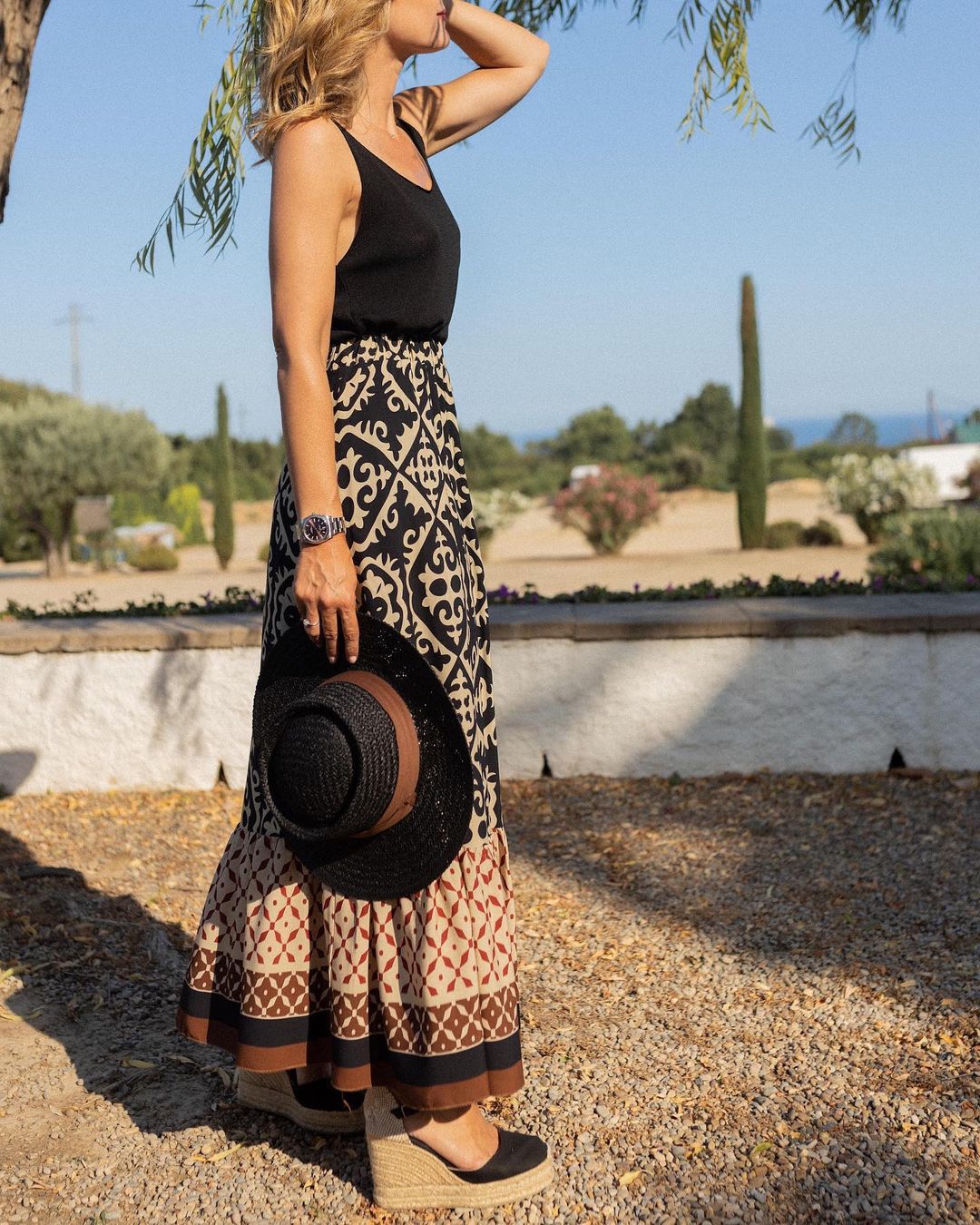 Femme en robe longue à motifs, portant un chapeau noir et des espadrilles compensées, dans un jardin méditerranéen ensoleillé.