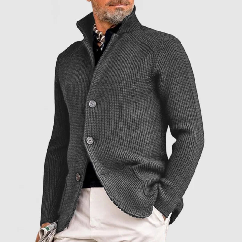 Homme portant un cardigan gris élégant, col relevé, boutons visibles, sur chemise noire et pantalon beige. Mode masculine chic et moderne.