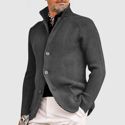 Homme portant un cardigan gris élégant, col relevé, boutons visibles, sur chemise noire et pantalon beige. Mode masculine chic et moderne.