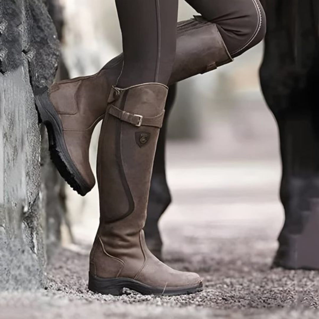 Bottes d'équitation marron en cuir, style équestre, portées par une personne en pantalon d'équitation, posées contre un mur en pierre.