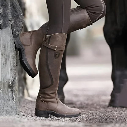 Bottes d'équitation marron en cuir, style équestre, portées par une personne en pantalon d'équitation, posées contre un mur en pierre.