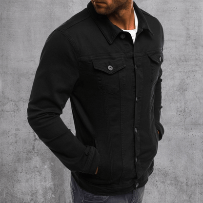 Homme portant une veste en jean noire, posant contre un mur gris. Mode masculine, style décontracté, veste tendance, tenue automne-hiver.