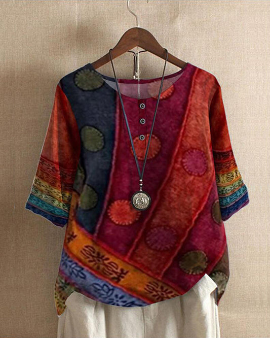 Blouse bohème colorée à motifs ethniques, manches trois-quarts, col rond avec boutons, suspendue sur un cintre en bois, fond beige.