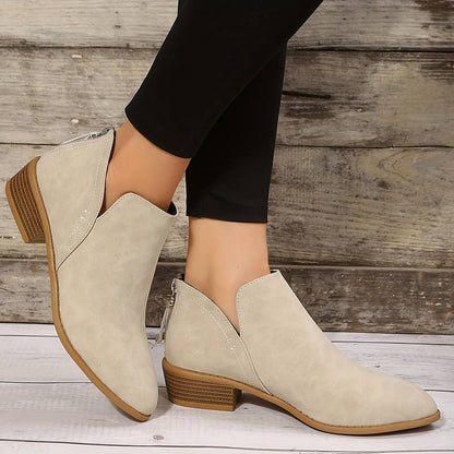 Bottines beiges en cuir synthétique, talon bas, fermeture éclair arrière, portées avec un pantalon noir, sur fond de bois rustique.