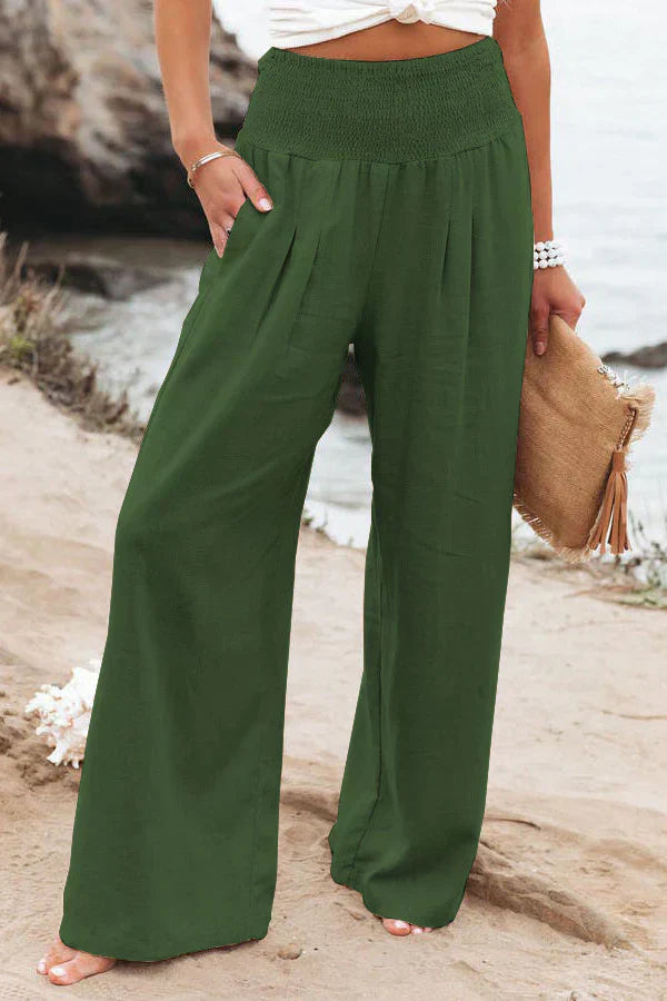 Pantalon large vert pour femme, taille haute, plage en arrière-plan, style décontracté, mode estivale, accessoire sac en paille, bord de mer.
