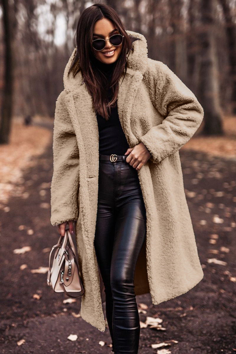 Femme portant un manteau en fausse fourrure beige, lunettes de soleil, pantalon en cuir noir, tenant un sac à main, marchant dans un parc automnal.