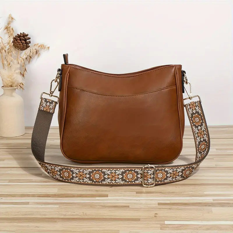 Sac à main en cuir marron avec bandoulière brodée florale, posé sur une table en bois clair, décor minimaliste avec vase et épis de blé.