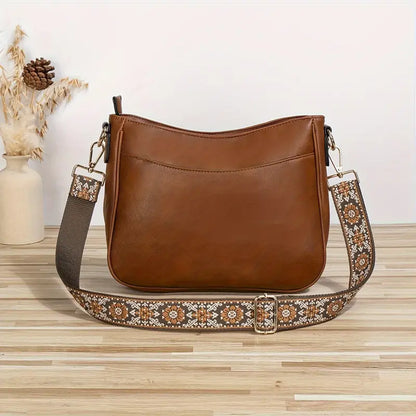 Sac à main en cuir marron avec bandoulière brodée florale, posé sur une table en bois clair, décor minimaliste avec vase et épis de blé.