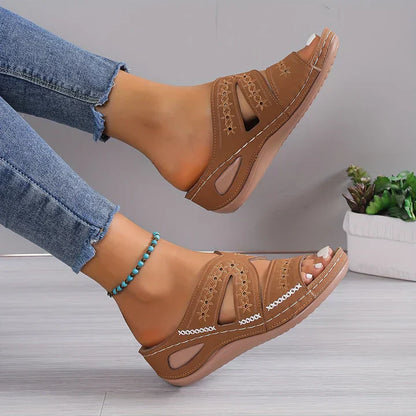 Sandales marron en cuir avec motifs ajourés portées avec un jean bleu, bracelet turquoise, posées sur un sol en bois, plante verte en arrière-plan.