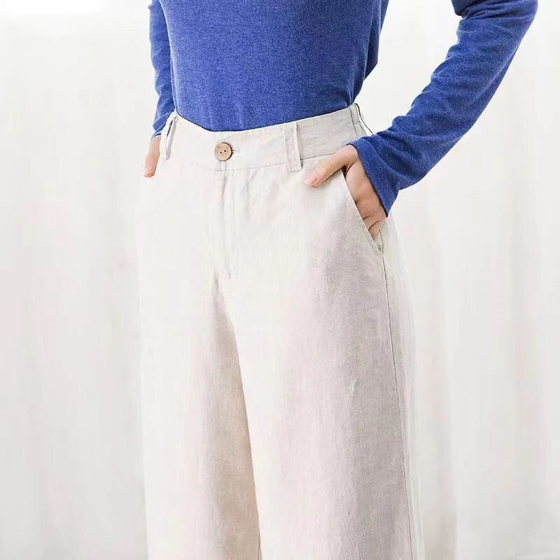 Personne portant un pantalon large beige en lin avec un bouton, mains dans les poches, et un pull bleu. Mode décontractée, style élégant et confortable.