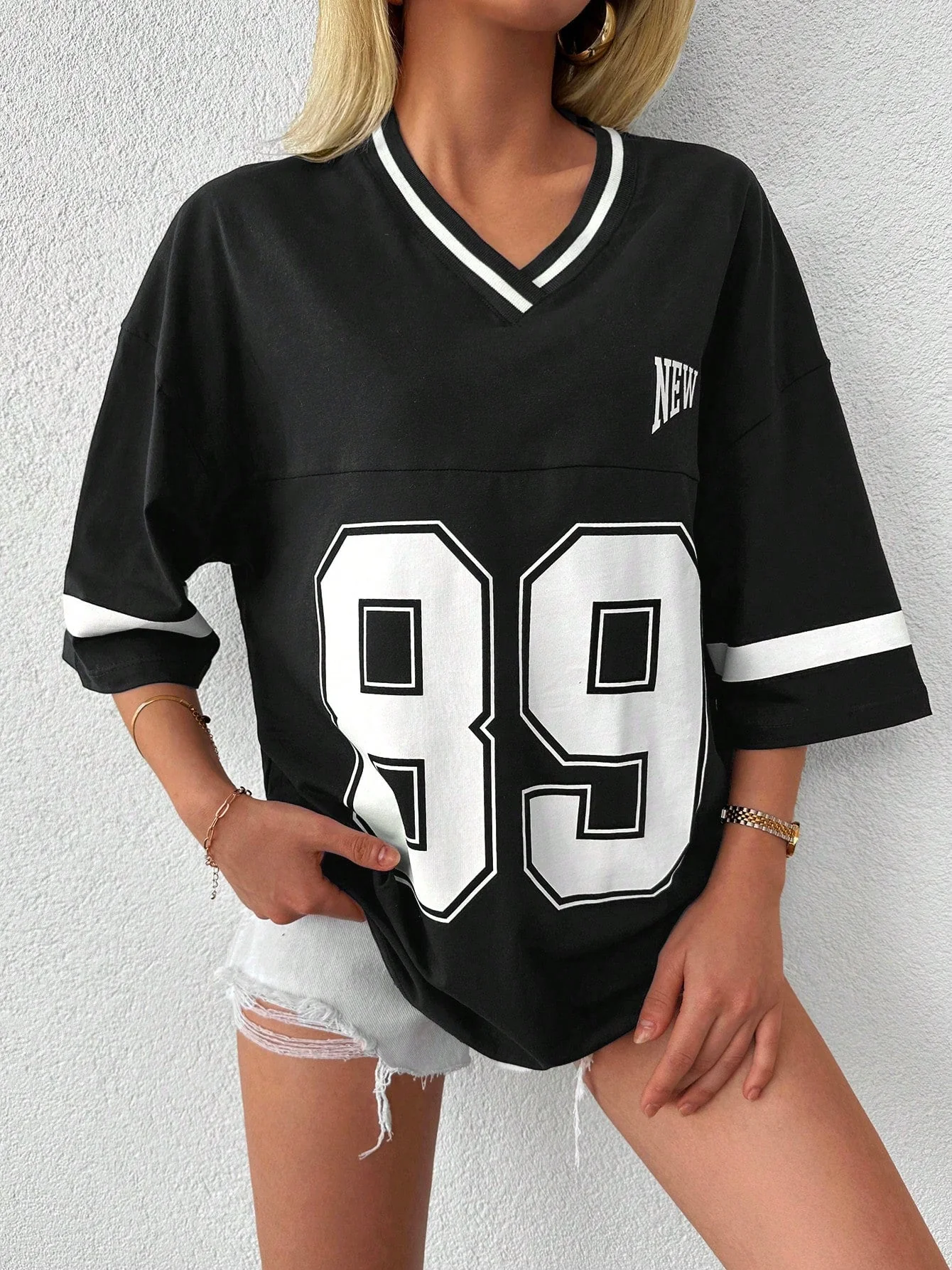 Femme portant un t-shirt noir à manches courtes avec numéro 89, style décontracté, sur short en jean déchiré. Mode urbaine, tendance sportive.