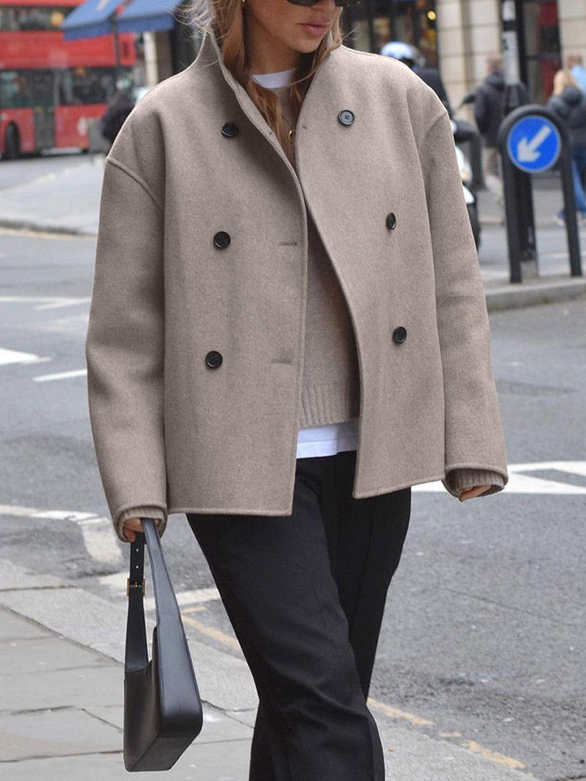Femme portant un manteau beige à double boutonnage, sac à main noir, rue urbaine, mode automne, style chic, bus rouge en arrière-plan.