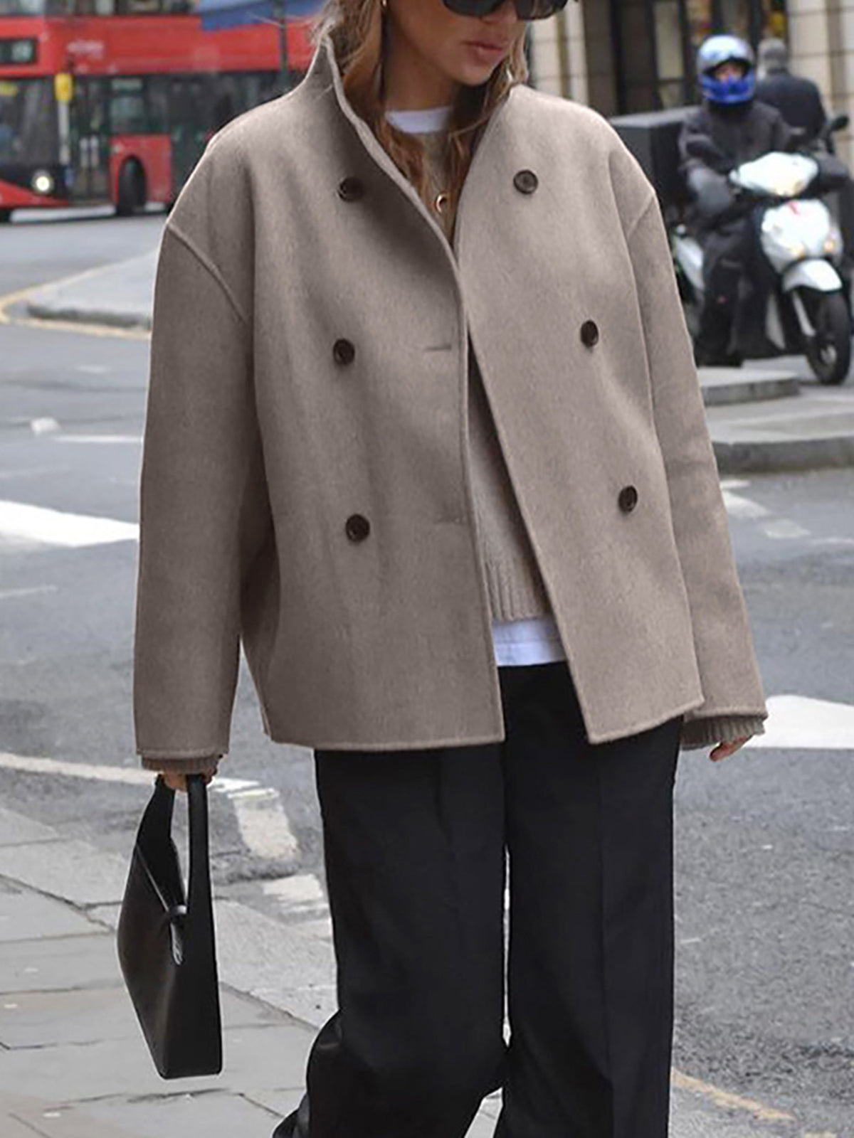 Femme portant un manteau beige à double boutonnage, pantalon noir, sac à main noir, traversant une rue urbaine avec bus rouge et scooter en arrière-plan.