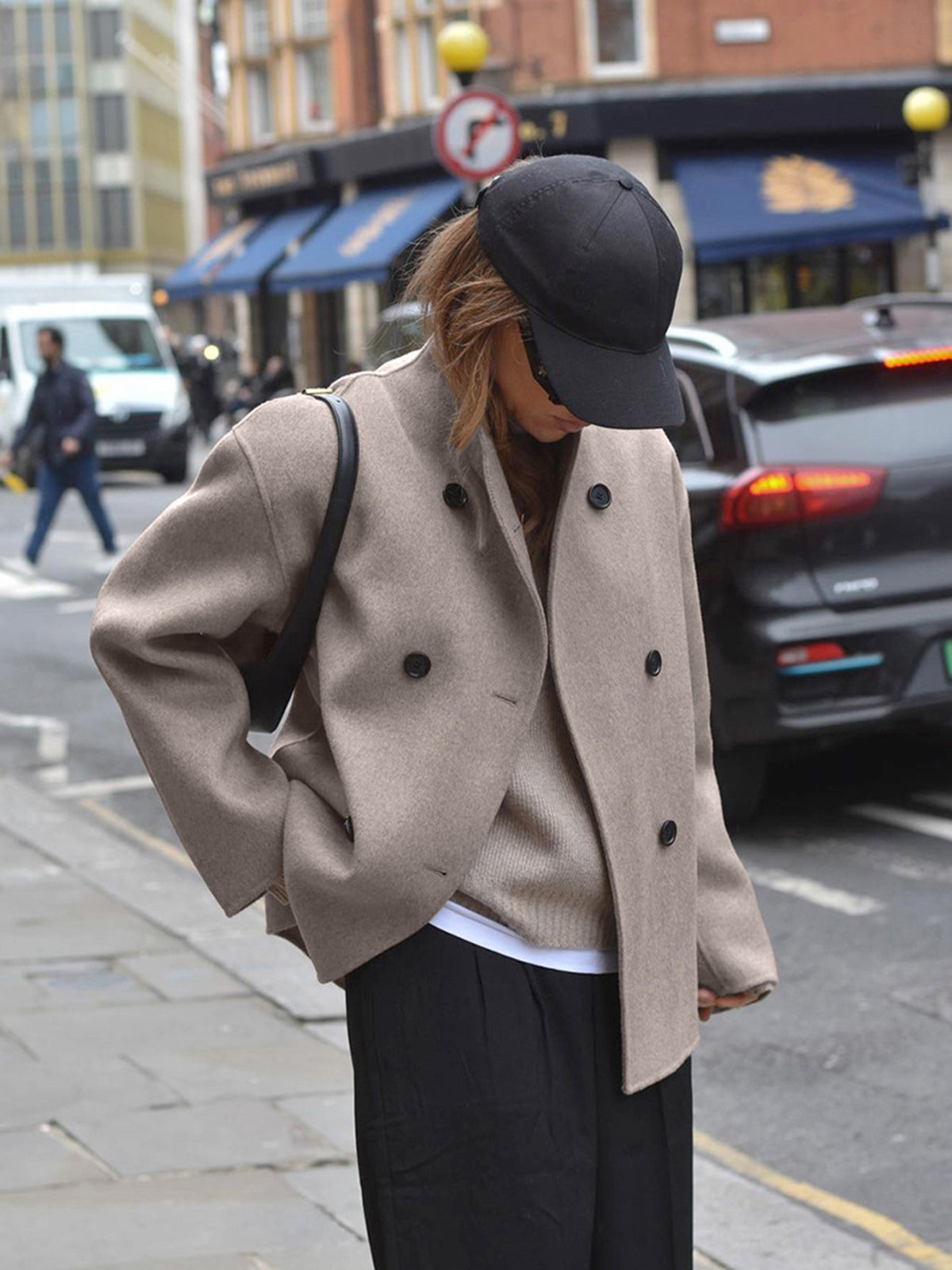 Femme portant un manteau beige, casquette noire, dans une rue urbaine. Mode automne, style décontracté, tendance vestimentaire urbaine.