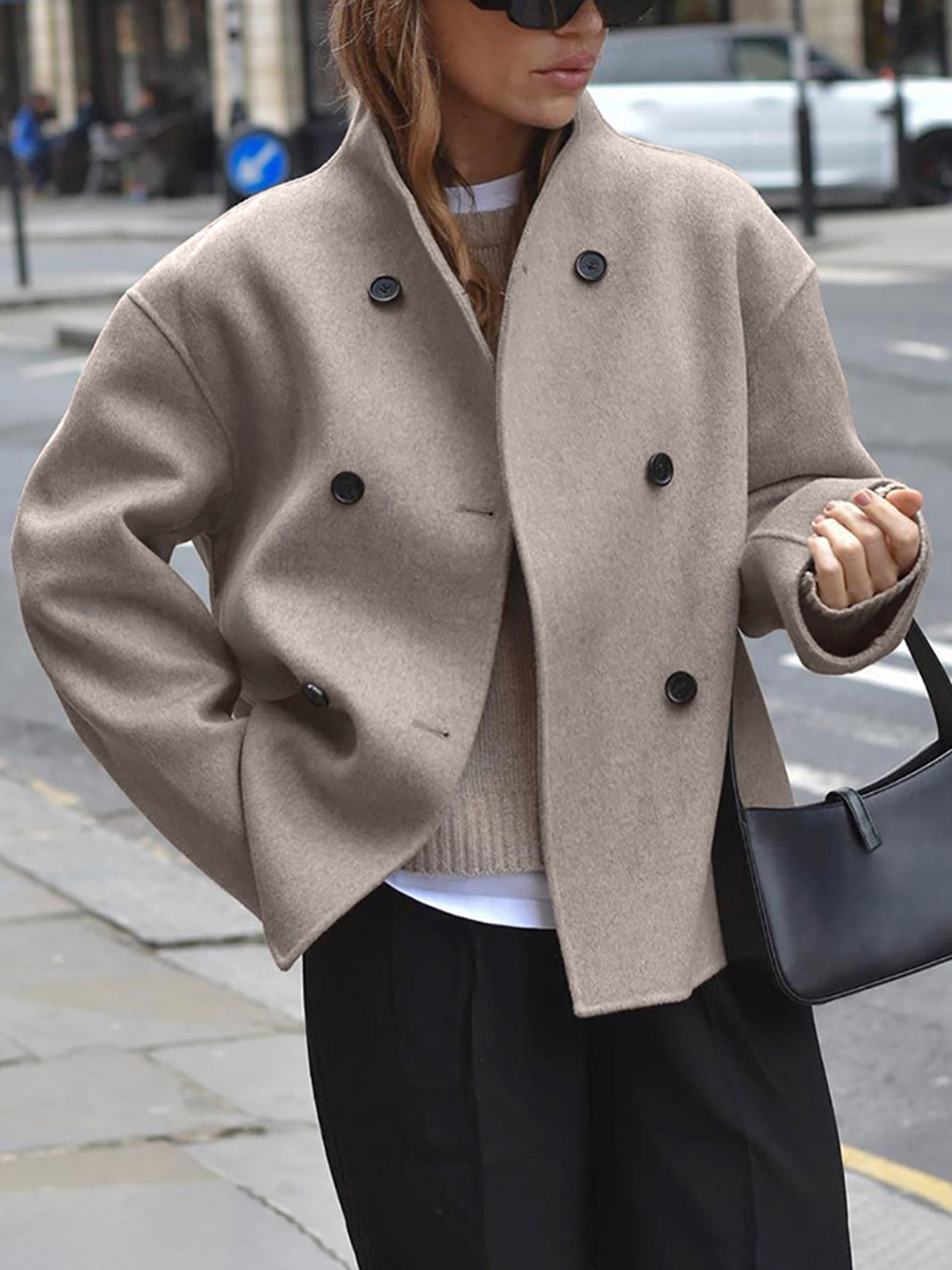 Femme portant un manteau beige à double boutonnage, lunettes de soleil noires, sac à main noir, debout dans une rue urbaine. Mode automne chic.
