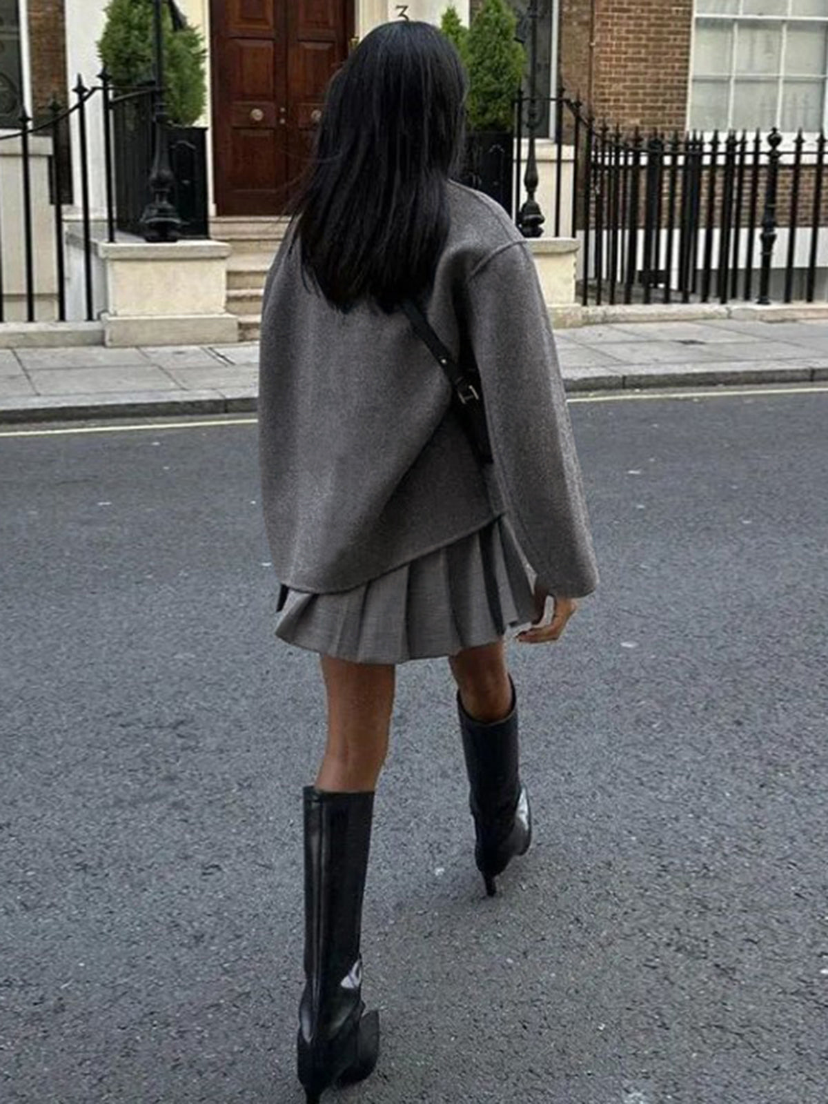 Femme en jupe plissée grise, pull oversize et bottes noires marchant dans une rue urbaine. Mode automne, style urbain chic.