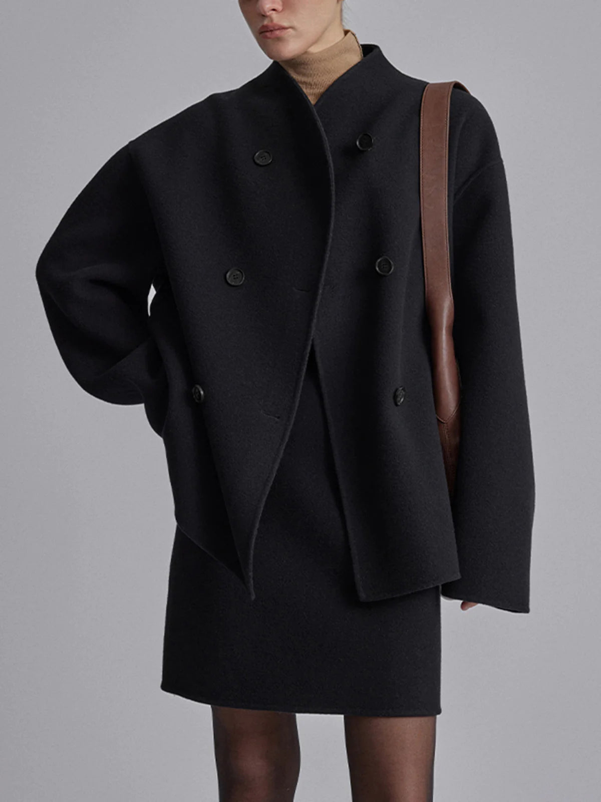 Femme portant un manteau noir oversize à double boutonnage, robe noire, col roulé beige, sac en cuir marron. Mode élégante et moderne.