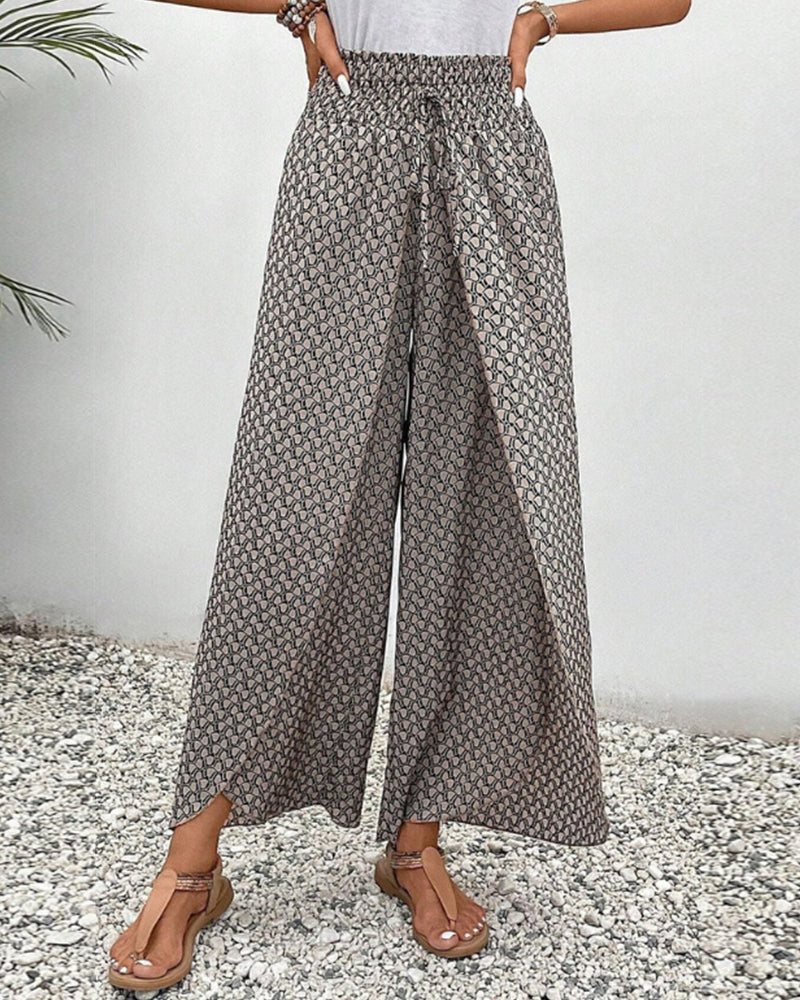 Pantalon large femme à motifs géométriques, taille haute, avec ceinture élastique, porté avec sandales marron sur fond de gravier et mur blanc.