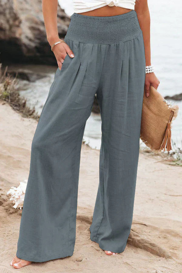 Pantalon large bleu-gris pour femme, taille haute, avec poches, porté sur une plage. Mode estivale décontractée, idéal pour un look chic et confortable.