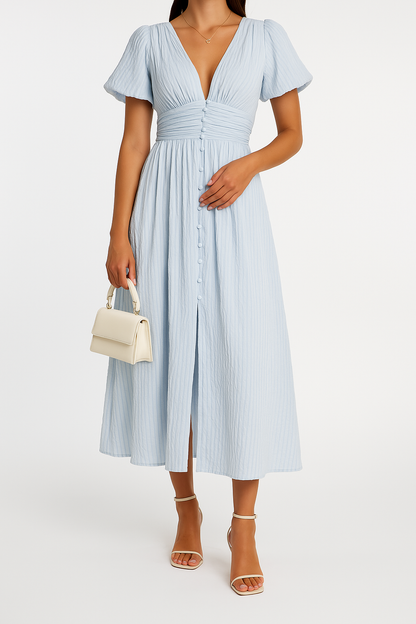 Robe longue bleue à manches courtes, décolleté en V, boutons devant. Femme tenant un sac à main blanc, portant des sandales à talons. Mode élégante.