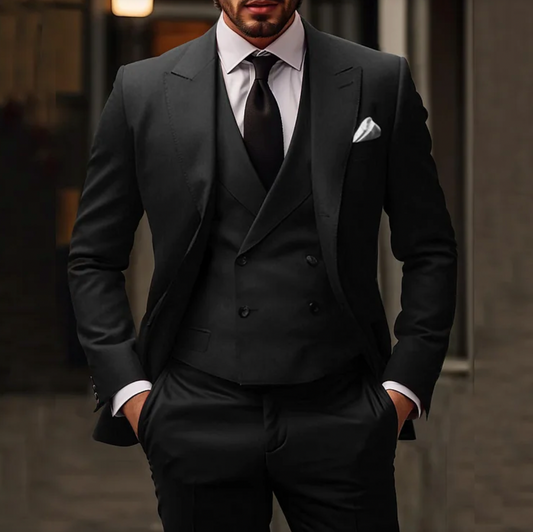 Homme en costume noir élégant, cravate noire, gilet, pochette blanche, mains dans les poches, style formel, mode masculine chic.