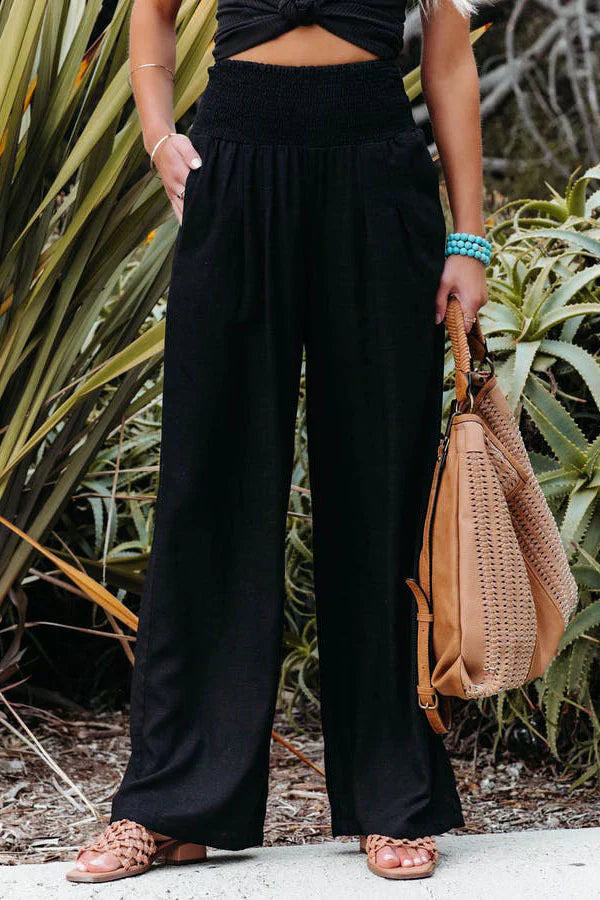 Femme en pantalon noir ample, tenant un sac en cuir beige, portant des sandales marron et un bracelet turquoise, posant devant des plantes vertes.