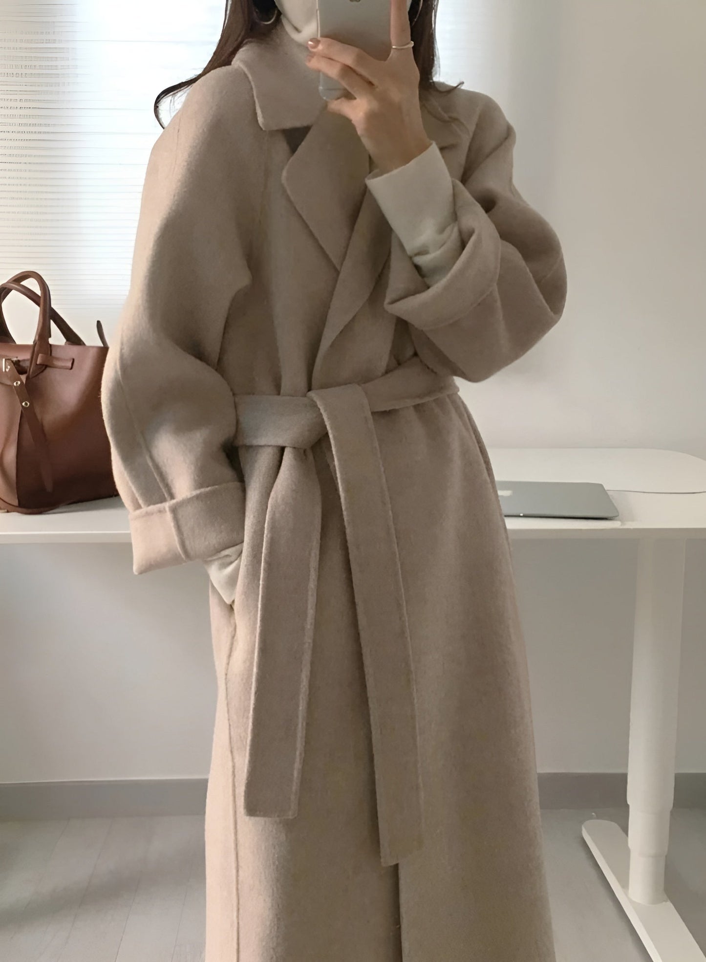 Manteau long beige ceinturé, élégant et moderne, porté par une personne dans une pièce lumineuse. Sac en cuir marron et ordinateur portable en arrière-plan.