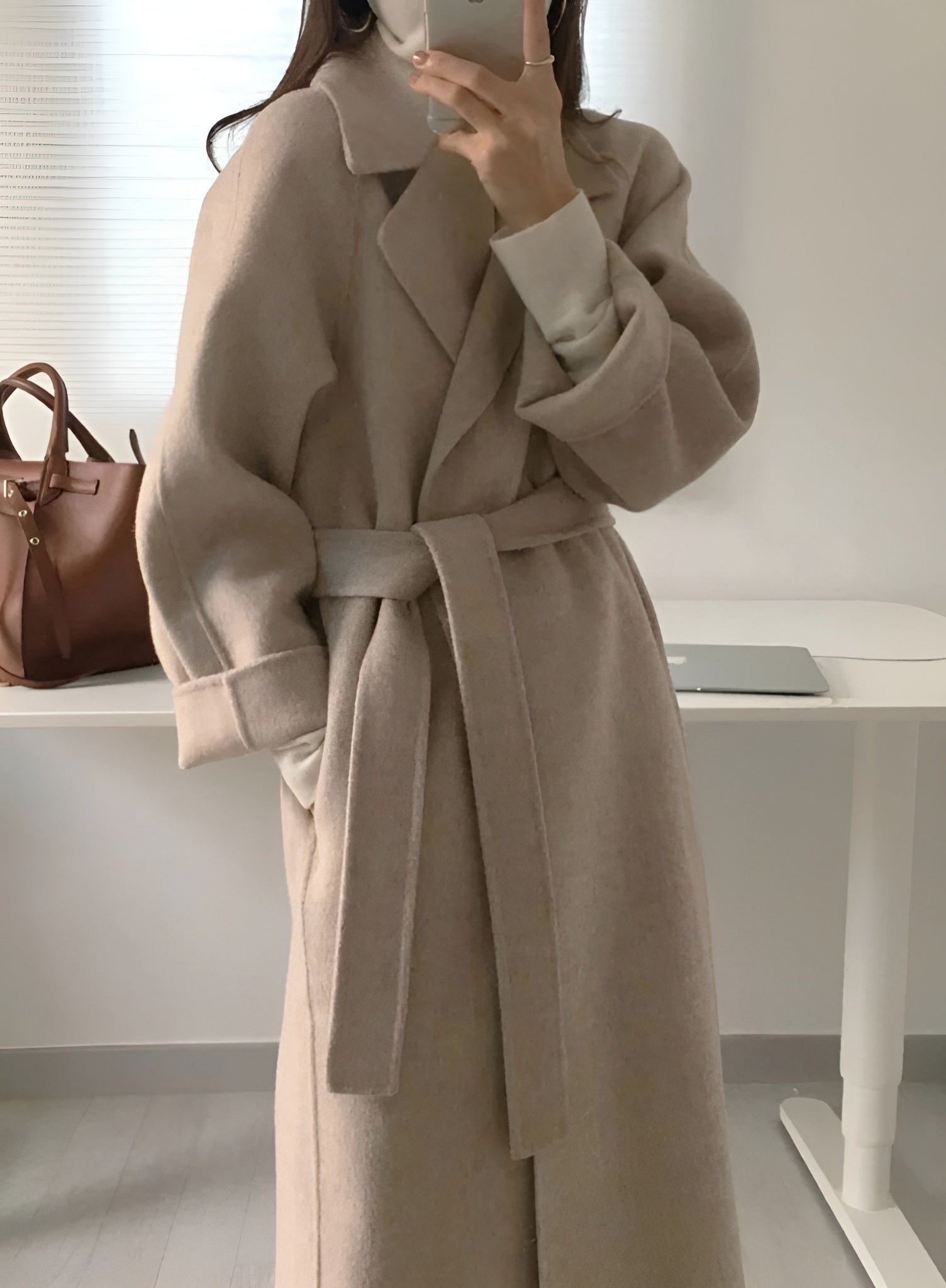 Manteau long beige ceinturé, élégant et moderne, porté par une personne dans une pièce lumineuse. Sac en cuir marron et ordinateur portable en arrière-plan.