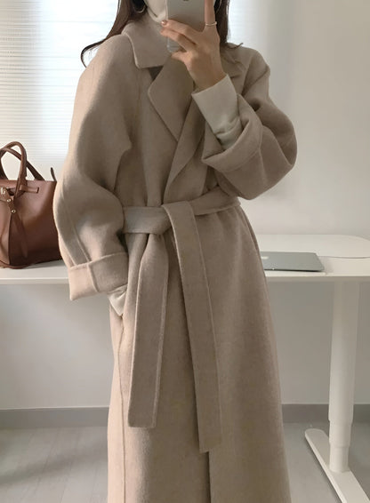 Manteau long beige ceinturé, élégant et moderne, porté par une personne dans une pièce lumineuse. Sac en cuir marron et ordinateur portable en arrière-plan.
