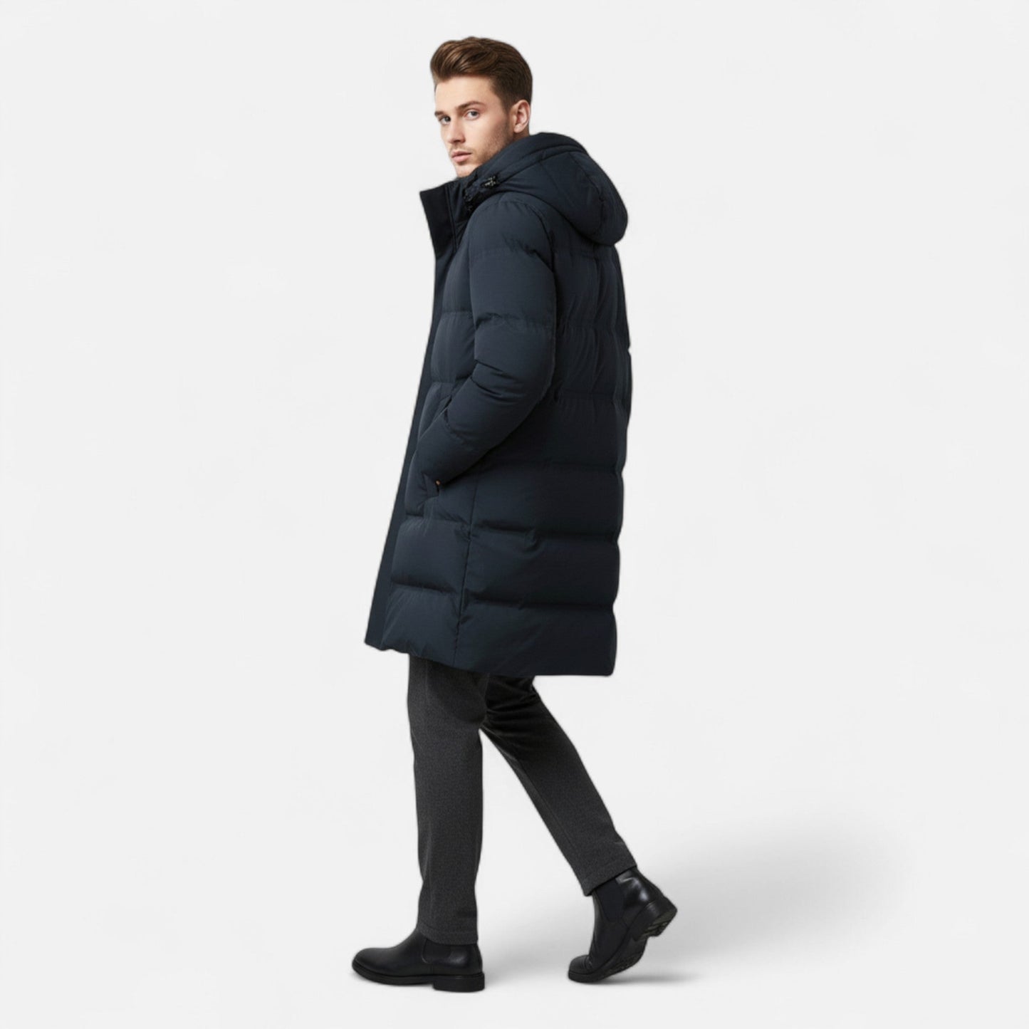 Homme portant un manteau d'hiver long et noir, style doudoune, avec capuche, marchant sur fond blanc. Mode masculine, vêtements chauds, hiver.