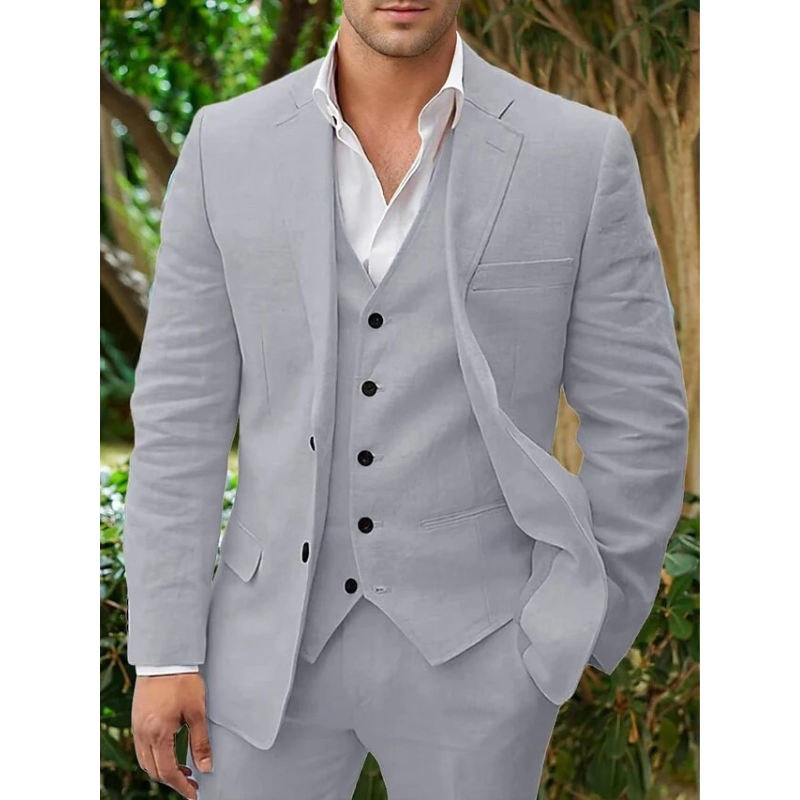 Homme en costume gris élégant, chemise blanche, cravate absente, posant devant un fond de verdure. Mode masculine, tenue formelle, style moderne.
