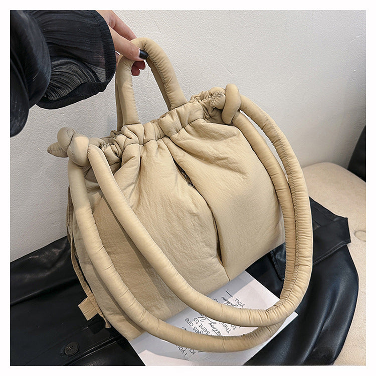 Sac à main beige en tissu plissé avec poignées doubles, design moderne et élégant, posé sur une surface noire. Accessoire mode tendance.