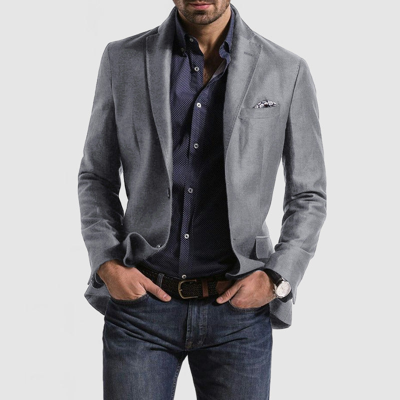 Homme portant une veste grise élégante, chemise bleue à motifs, jean décontracté, montre-bracelet et pochette, style mode masculine moderne.