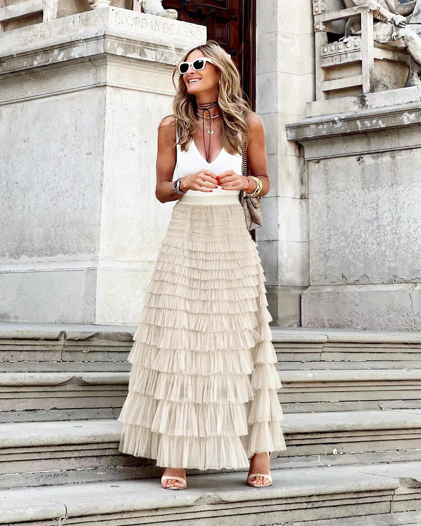 Femme élégante en jupe longue à volants beige et top blanc, posant sur des marches en pierre, lunettes de soleil blanches, style chic urbain.