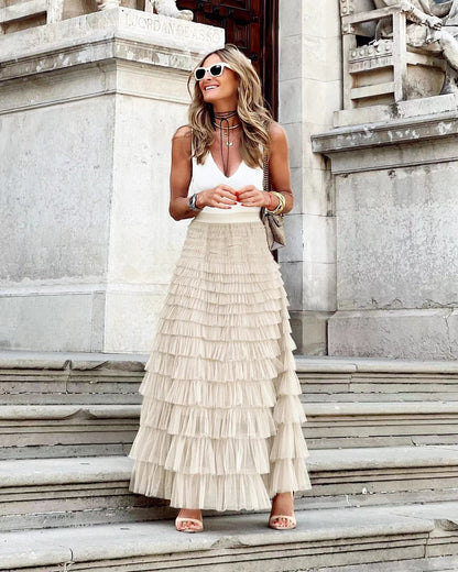 Femme élégante en jupe longue à volants beige et top blanc, posant sur des marches en pierre, lunettes de soleil blanches, style chic urbain.