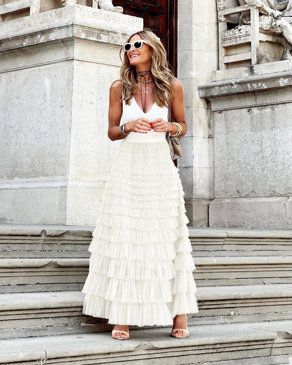 Femme élégante en jupe longue plissée blanche et débardeur blanc, posant sur des marches en pierre, devant un bâtiment historique. Mode chic et estivale.