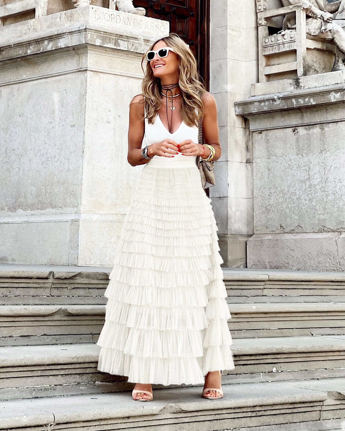 Femme élégante en jupe longue plissée blanche et débardeur blanc, posant sur des marches en pierre, lunettes de soleil rondes, style chic urbain.