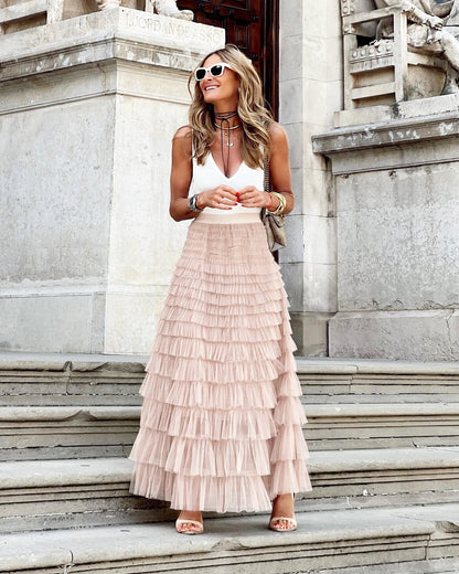 Femme souriante en jupe longue à volants rose et débardeur blanc, posant sur des marches en pierre, lunettes de soleil blanches, style chic et décontracté.