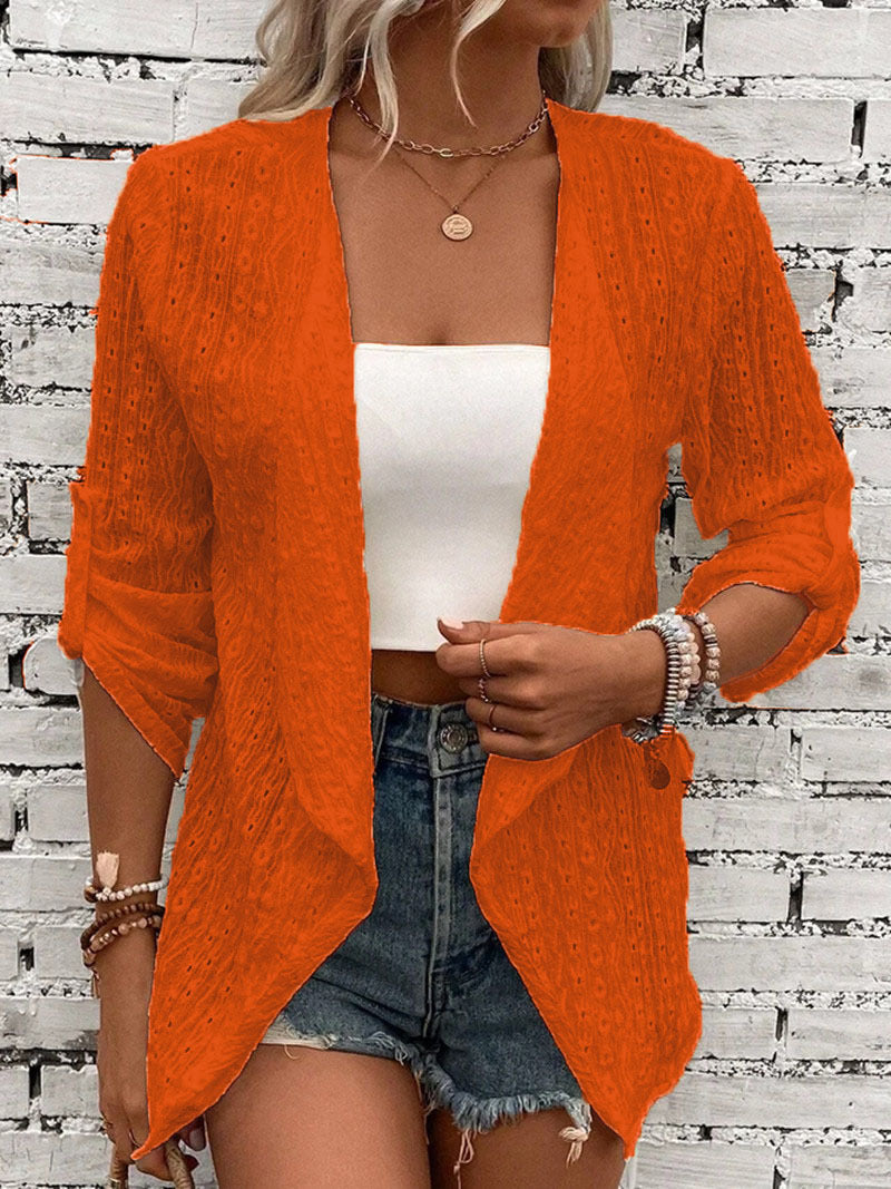 Femme portant un cardigan orange en dentelle, débardeur blanc et short en jean devant un mur de briques blanches. Mode estivale décontractée.
