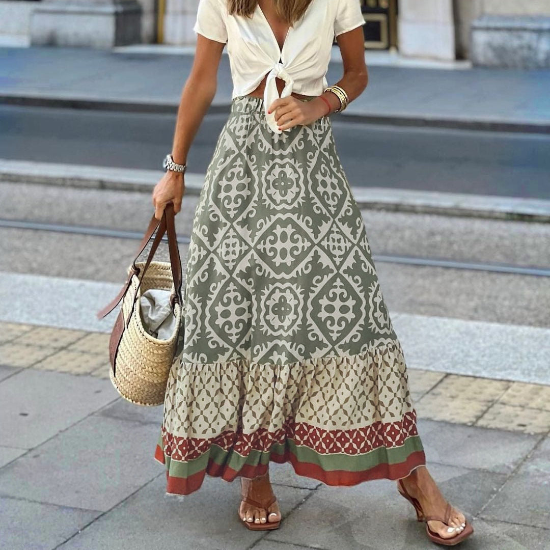 Femme en jupe longue bohème à motifs géométriques, haut blanc noué, sandales, sac en osier, marchant en ville. Mode estivale décontractée.