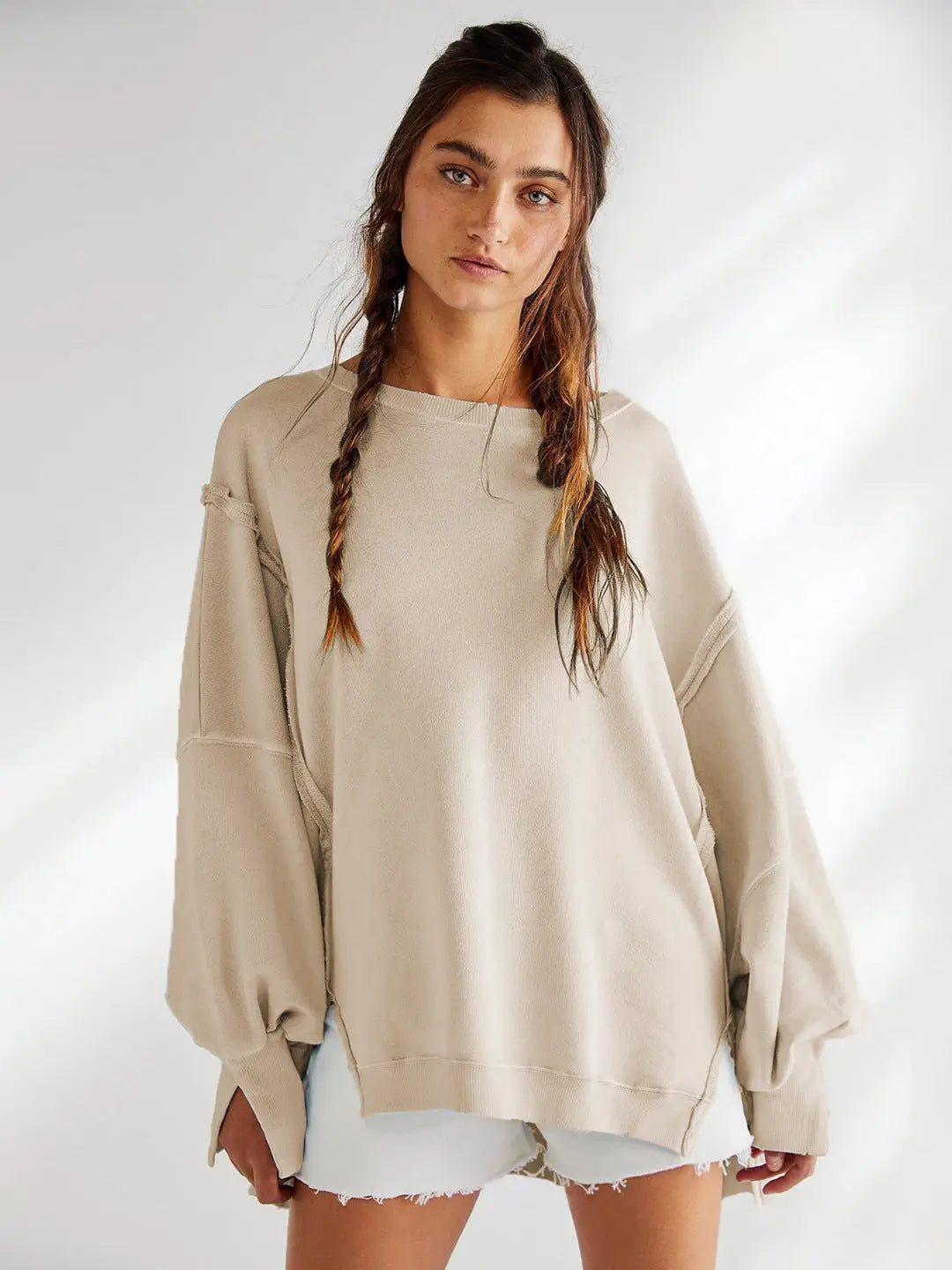 Jeune femme portant un pull beige ample avec manches longues, cheveux tressés, posant devant un fond blanc. Mode décontractée, style automne-hiver.