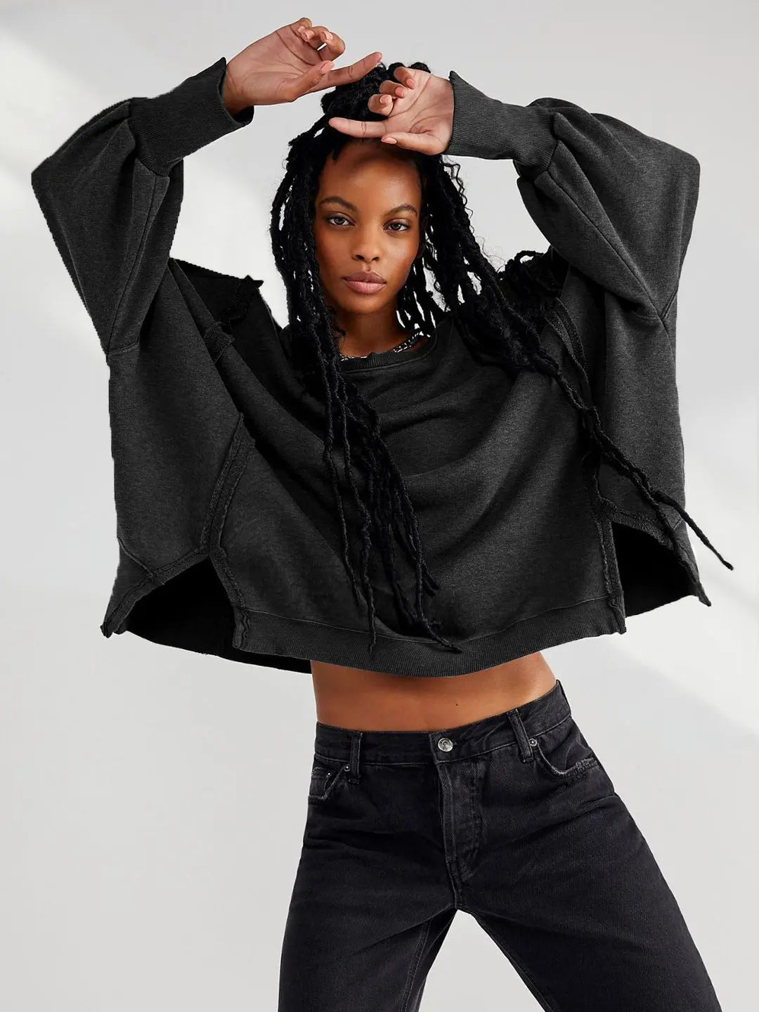 Jeune femme portant un sweat-shirt noir ample et un jean foncé, posant avec les bras levés. Mode décontractée, style urbain, cheveux tressés.