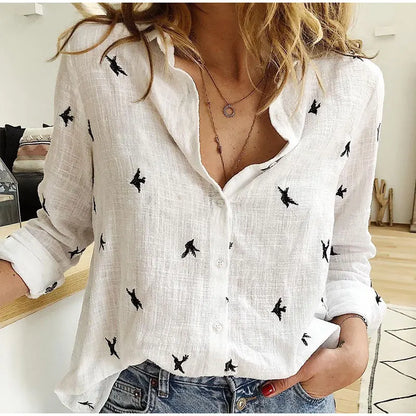 Chemise blanche en lin avec motifs d'oiseaux noirs, manches retroussées, portée par une femme. Style décontracté, tendance mode féminine.